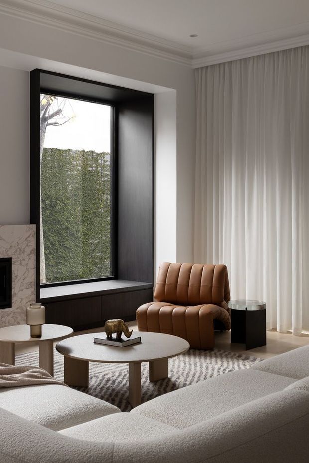 A refined contemporary living room with a large black-framed window, dark built-in window seat cabinetry, sculptural brown upholstered lounge chair, rounded coffee tables, a textured neutral rug, and a softly curved cream sectional sofa. Floor-to-ceiling sheer curtains, a marble fireplace surround, and detailed ceiling cornices create a warm, sophisticated interior.