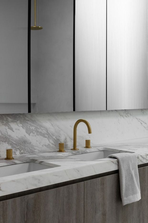 Close-up of a refined bathroom vanity featuring a double integrated basin set into a heavily veined white marble countertop and backsplash. The vanity has vertical-grain dark timber cabinetry, brushed brass/gold tapware, and a large mirror with vertically ribbed detailing. A neutral grey hand towel hangs over the counter edge.
