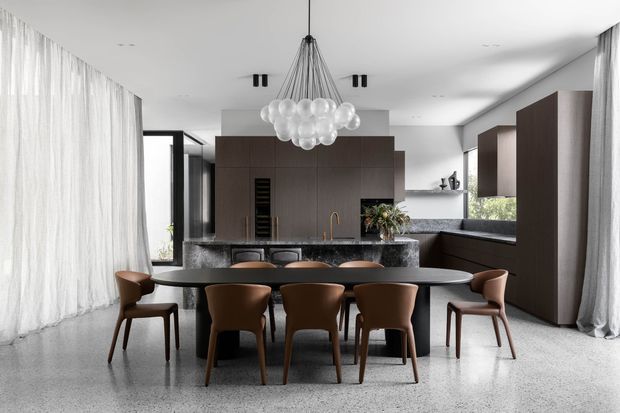 A refined open-plan dining and kitchen interior with a long dark dining table surrounded by leather dining chairs. The space features dark timber cabinetry, a grey stone kitchen island, polished terrazzo flooring, sheer full-height curtains, large glazed openings, and a sculptural cluster pendant light.