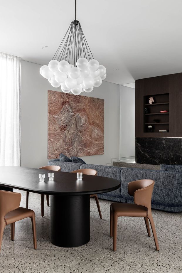 A refined contemporary open-plan dining and living area with a large oval black dining table, tan upholstered dining chairs, a textured blue-grey sofa, and a sculptural cluster pendant light made of translucent white globes. The room features a light terrazzo floor, white walls and sheer curtains, dark timber built-in shelving, a dramatic black marble fireplace surround, and a large abstract textured artwork.