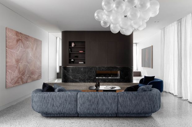 A refined contemporary living room centered on a large curved blue-grey sectional sofa facing a dark marble fireplace wall with integrated dark timber cabinetry and shelving. A sculptural cluster pendant hangs overhead, while sheer floor-to-ceiling curtains, terrazzo-style flooring, neutral walls, and abstract artwork create a polished, calm interior.