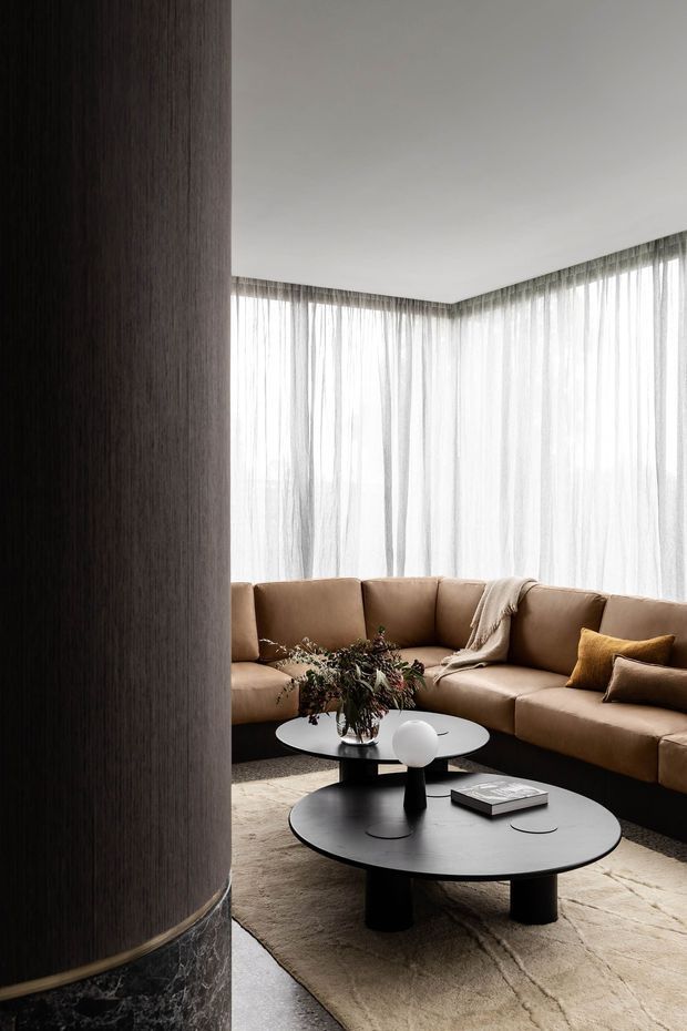 A refined living room viewed past a tall dark timber-clad column with a marble base. The space features a large tan sectional sofa, sheer full-height curtains wrapping around corner glazing, layered round black coffee tables, a neutral textured rug, and restrained decorative accents. The overall look is warm, sophisticated, and contemporary.