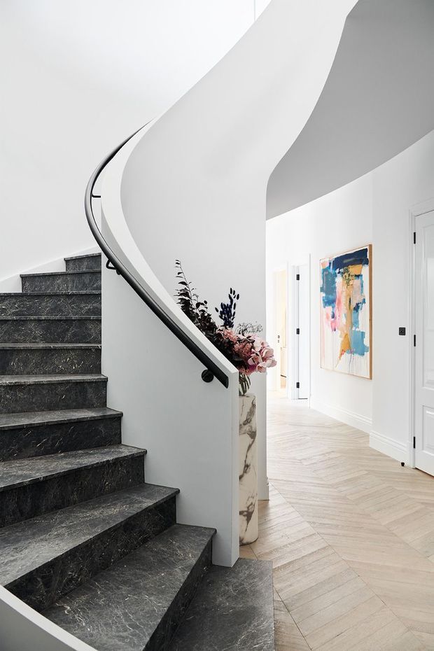 A refined residential stairway with a dramatic curved white balustrade and dark veined stone stair treads. The bright hallway beyond has white paneled doors, pale herringbone timber flooring, a marble-look pedestal with floral arrangement, and colourful abstract artwork. The composition emphasizes the sweeping sculptural staircase and high-end monochromatic interior.