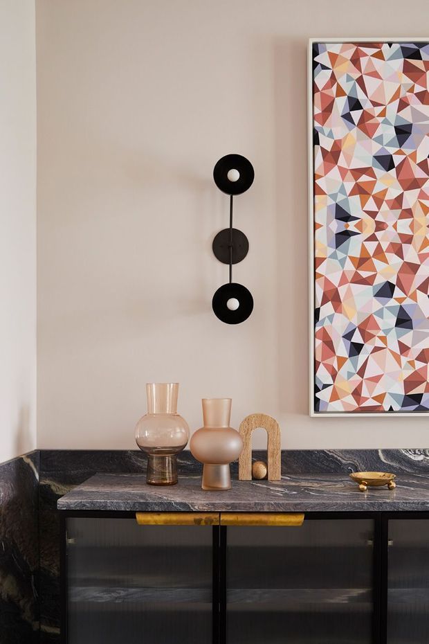 A refined interior vignette featuring a dark marble-topped sideboard with black cabinetry, set against a warm beige wall. A sculptural black double wall sconce, colourful geometric framed artwork, glass and ceramic vessels, and a small brass decorative object create a sophisticated, art-led composition.
