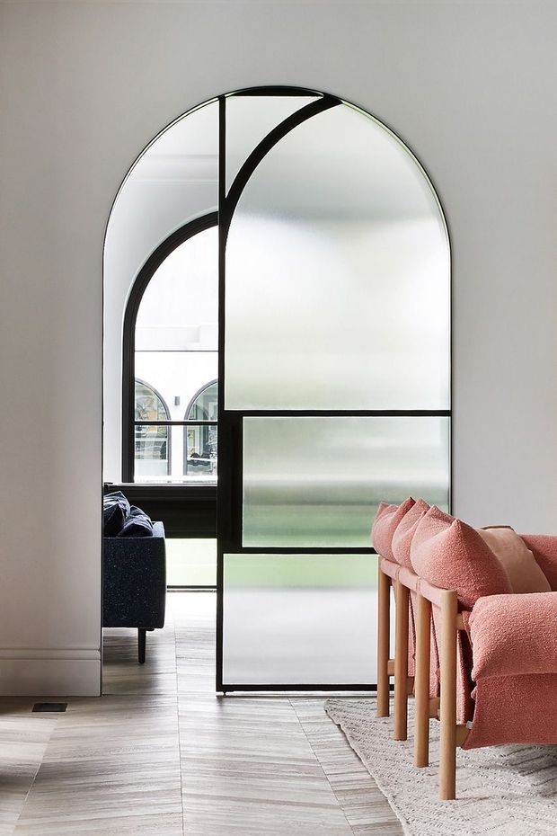 A refined interior featuring a tall arched black-framed glazed partition or door separating a light living area from an adjoining room. The space has pale timber flooring, white walls, a textured neutral rug, and a row of sculptural blush-pink upholstered dining or lounge chairs with light timber legs. Reflections reveal an adjacent dark sofa and another arched window, while the overall composition feels contemporary and graphic.