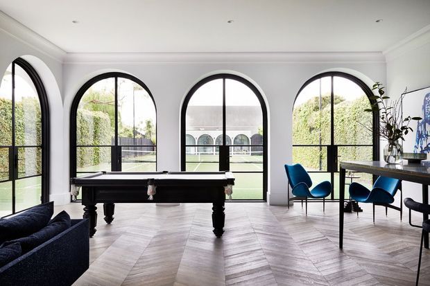 Elegant light-filled living and entertaining room with a black billiards table, sculptural blue lounge chairs, a dark dining or console table, and dramatic arched steel-framed windows overlooking a courtyard. The room features pale herringbone timber flooring, white walls with decorative cornice moulding, and a refined contemporary-classic aesthetic.