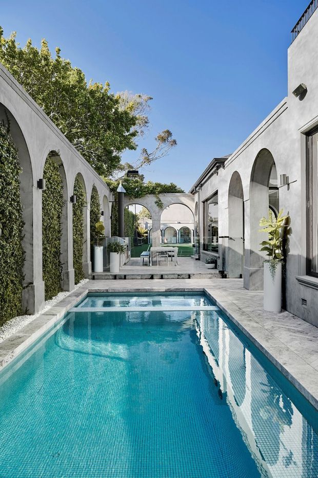 A refined outdoor courtyard centered on a long turquoise swimming pool, enclosed by pale rendered walls with repeated arched openings and climbing greenery. Light stone paving, contemporary glazing, sculptural planters, and a small outdoor seating area create a calm, resort-like architectural setting under a clear blue sky.