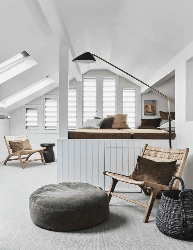 Bright, airy attic-style living space with a raised built-in daybed or seating platform beneath a sloped white ceiling. The room features extensive white-framed windows with roller blinds, pale flooring, timber-and-cane lounge chairs, a large dark round ottoman, woven basket, black accent table, and a sculptural arched floor lamp. The restrained palette of white, beige, taupe, and charcoal creates a calm, contemporary coastal atmosphere.