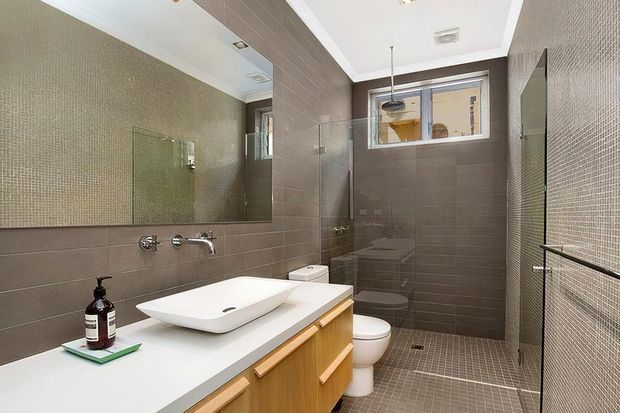 Contemporary bathroom with a wall-mounted basin on a timber vanity, large mirror, wall-hung fixtures, toilet, frameless glass shower enclosure, and small-format grey mosaic wall and floor tiles. A high horizontal window provides natural light.