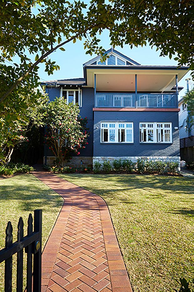 Front exterior view of a two-storey brick house with a dark painted brick facade, white-framed windows, upper-level covered balcony, and a prominent gabled roof. A curved herringbone brick walkway leads through the front lawn to the entrance, with mature landscaping framing the property.
