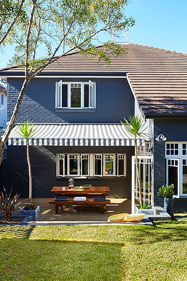 Exterior view of a dark-painted, two-storey residential house with a tiled pitched roof, white-trimmed windows, and a striped retractable awning over a covered patio. The outdoor area includes a timber picnic-style dining table, potted plants, lawn, and a glazed side enclosure.