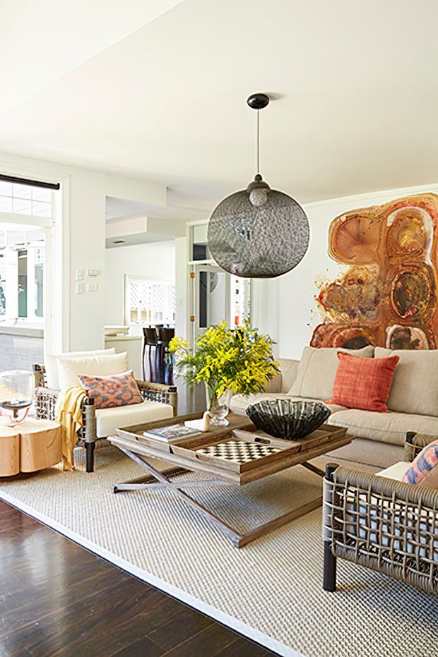 Bright, art-filled living room with a large cream sectional sofa, woven armchairs, a substantial wood-and-metal coffee table, patterned cushions, and a sculptural pendant light. A large abstract artwork forms a feature wall, while dark timber flooring and a textured neutral rug provide contrast. The open-plan background includes glimpses of a dining area and kitchen.