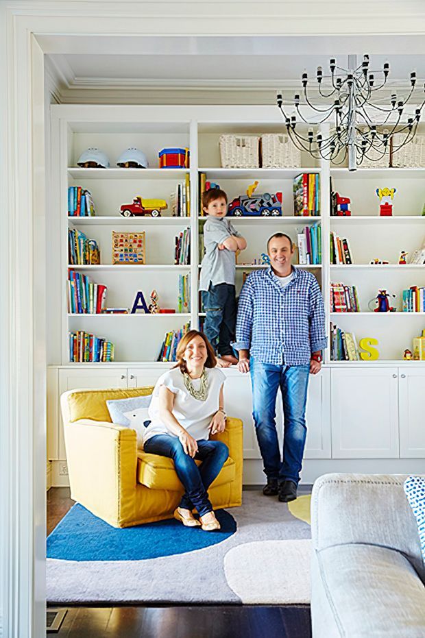 Bright family living room with extensive built-in white shelving and lower cabinetry filled with books, toys, and baskets. A yellow armchair, pale sectional sofa, layered blue and grey rugs, and a prominent black multi-arm chandelier create a cheerful, family-oriented interior.