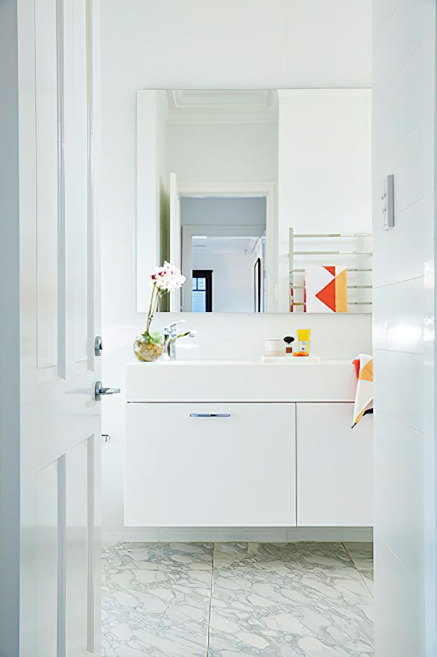 Bright, compact white bathroom viewed through an open door, featuring a floating white vanity with integrated basin, chrome tapware, large rectangular mirror, open wall shelving, white tiled walls, and veined marble-look floor tiles. Small orange and red accents appear in toiletries and towels.