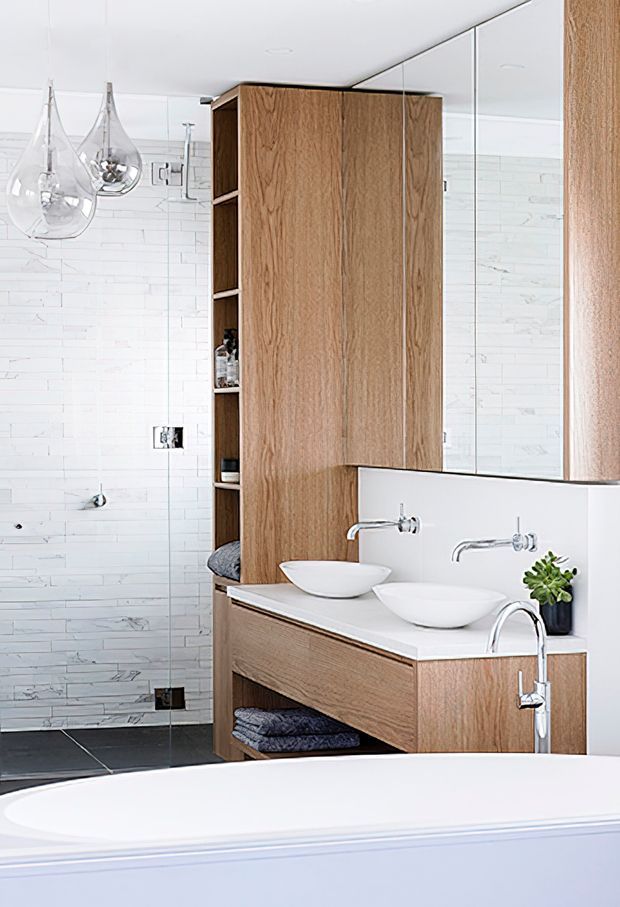 Bright contemporary bathroom with a freestanding bathtub in the foreground, a double floating timber-veneer vanity with two white vessel basins, wall-mounted chrome taps, mirrored cabinetry, open shelving, and white rectangular wall tiles. Dark floor tiles and sculptural clear pendant lights provide contrast.