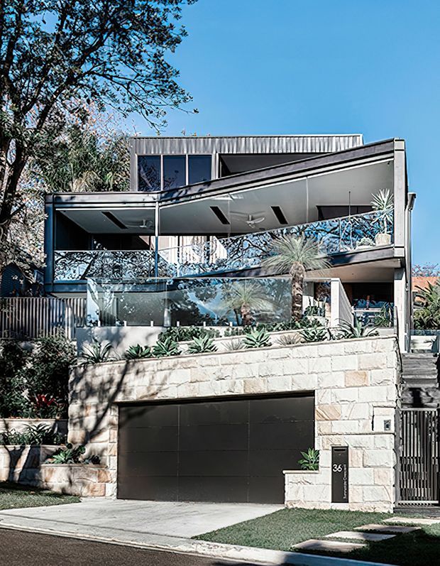 Street-facing view of a contemporary multi-level urban residence with a strong sloping roofline, projecting upper balcony, extensive glass balustrades, and a pale stone-clad lower level containing a dark garage door. The facade is integrated with landscaped terraces and established vegetation.