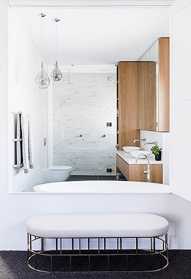 Bright contemporary bathroom viewed from the foreground, with a freestanding white bathtub, large wall mirror, wall-mounted toilet, walk-in shower, white marble-look stacked tile wall, and warm timber vanity cabinetry. Chrome fixtures, clear pendant lights, and a soft upholstered bench over a dark textured floor create a clean, refined contrast.