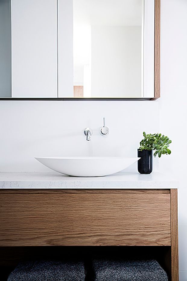 Minimalist bathroom vanity featuring a white vessel basin, wall-mounted chrome tapware, pale stone benchtop, and a warm timber floating vanity with open shelving. A mirrored cabinet and small potted plant complete the clean, refined composition.