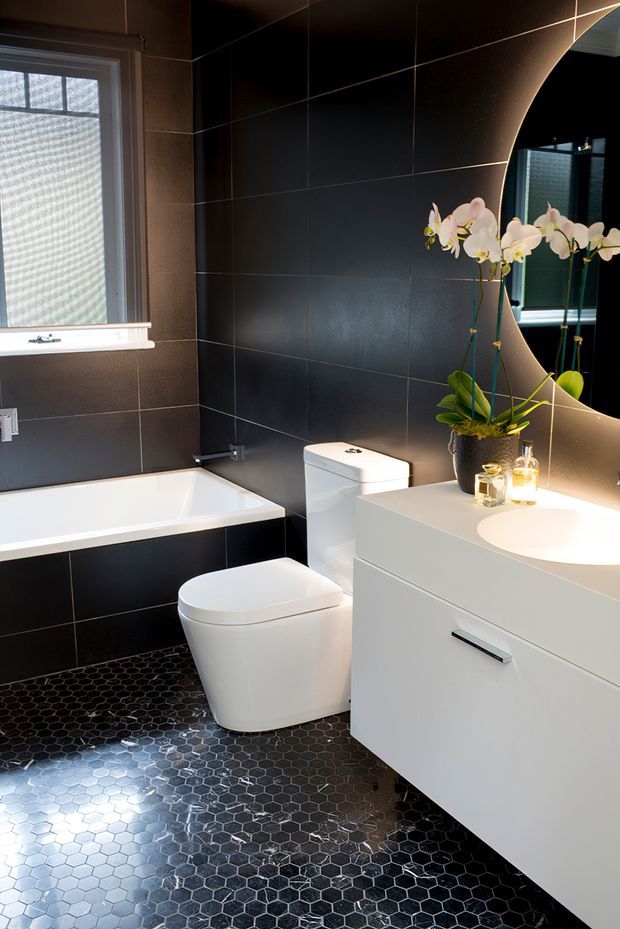 A compact contemporary bathroom featuring dark large-format wall tiles, black hexagonal floor tiles, a white bathtub, wall-mounted white vanity with integrated basin, and a modern close-coupled toilet. A large curved-edge mirror, orchids, and warm lighting add contrast and softness.