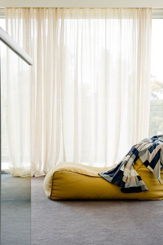 Bright contemporary interior corner featuring floor-to-ceiling sheer cream curtains, expansive glazing, grey carpet, and a low mustard-yellow upholstered lounge or beanbag with a geometric blue, cream, and mustard throw. A glass balustrade or partition is visible at left, with soft daylight creating an airy, relaxed atmosphere.