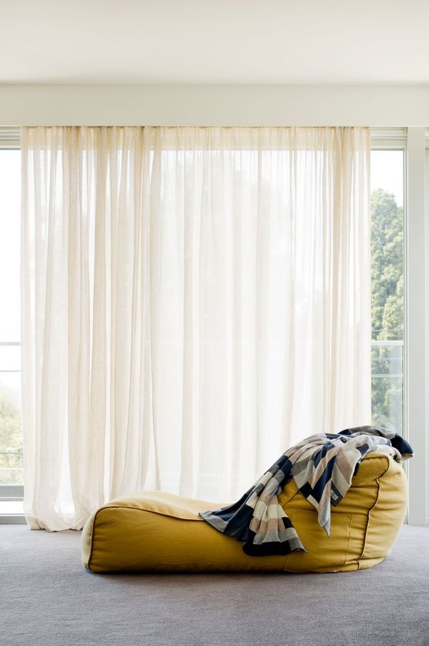 Bright, minimal interior featuring a low mustard-yellow upholstered lounge chair with a draped patterned throw, positioned on a light grey carpet in front of full-height windows with sheer white curtains. The room has abundant natural light and a calm, airy atmosphere.