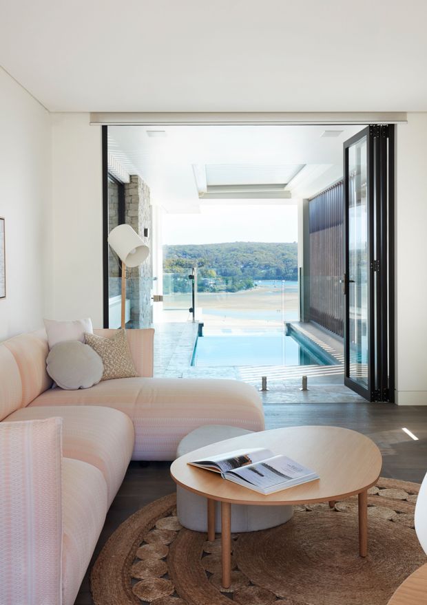 Bright contemporary living room opening directly onto a covered outdoor pool terrace, with expansive water views beyond. The room features a pale blush sectional sofa, round timber coffee table, textured round rug, exposed stone wall, dark timber or aluminium-framed folding doors, and a skylit white ceiling. A narrow swimming pool and glass balustrade extend toward the landscape.