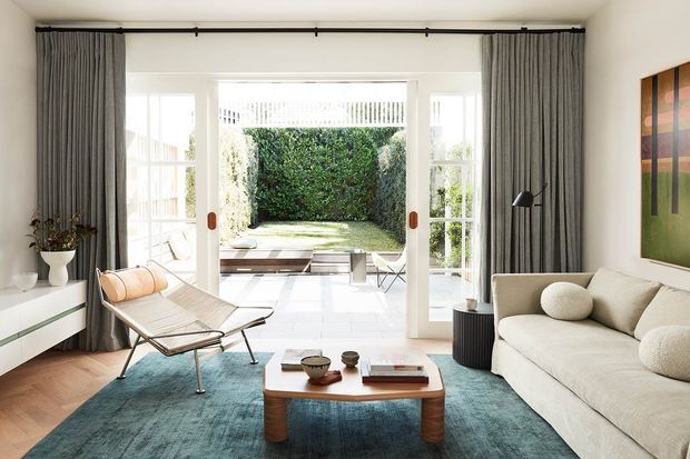 Bright contemporary living room with floor-to-ceiling glazed doors opening to a landscaped courtyard. The room features a cream sofa, sculptural lounge chair, walnut-toned coffee table, teal area rug, dark grey full-height curtains, white built-in cabinetry, and a black floor lamp. A colourful abstract artwork adds a strong wall accent.