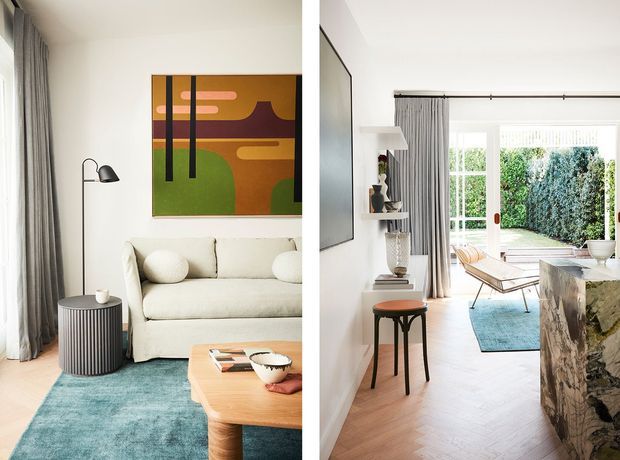 Bright, refined living room shown from two complementary angles, with an off-white sofa, turquoise rug, pale timber coffee table, abstract artwork, sculptural floor lamp, grey curtains, and a large glazed opening to a leafy garden. The room continues into a light timber-floored open-plan area with a small side table, shelving, dining or lounge furniture, and a rustic stone-clad kitchen island or feature element.