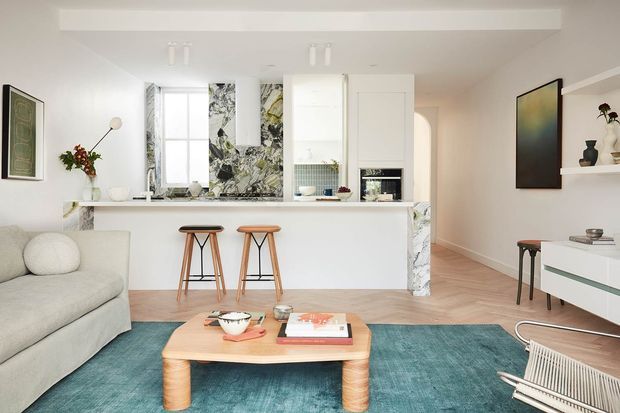 Bright open-plan living room and kitchen with a white minimalist interior, marble-patterned feature surfaces around the kitchen window and fireplace, light timber flooring, a compact island with two stools, a pale sofa, teal rug, and mid-century-inspired timber coffee table. Built-in shelving, artwork, and contemporary furniture add restrained character.