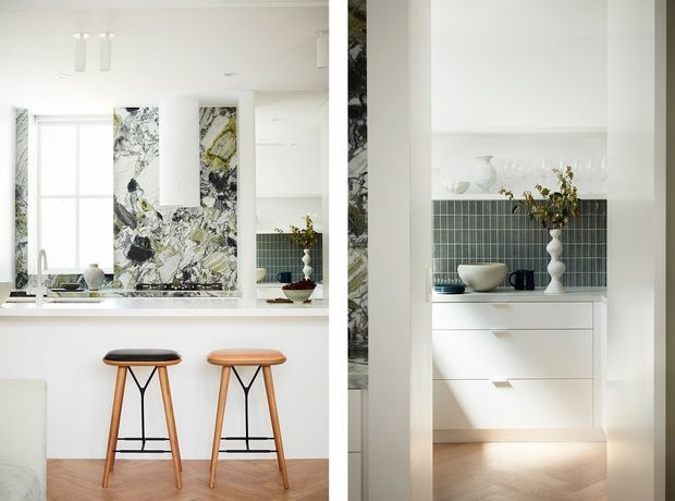 Bright, refined white kitchen with a compact island or breakfast bar, two timber stools, pale timber flooring, and a striking marble-patterned splashback. A built-in white storage and display unit with a dark green vertical-tile splashback is visible at right, styled with ceramics and glassware. The space is lit by generous natural light and minimalist pendant fixtures.