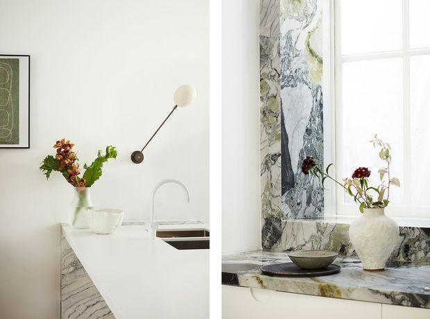 Bright, refined kitchen interior featuring a white island with integrated sink and sculptural curved faucet, white cabinetry, and dramatic veined marble used for the backsplash, counter edges, and window ledge. A large divided window provides abundant natural light, while sculptural floral arrangements, ceramic vessels, and a minimalist wall sconce add decorative detail.