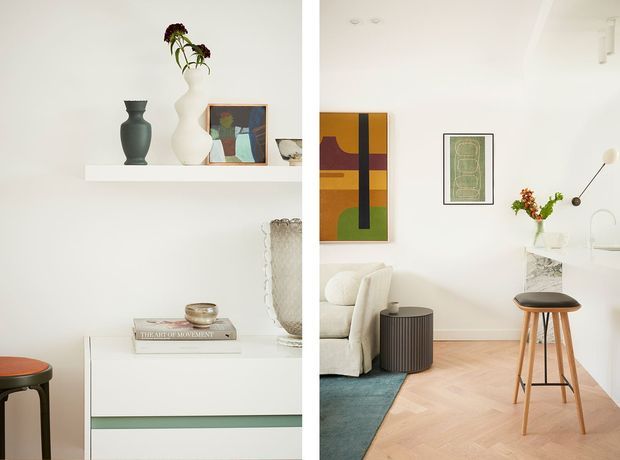 Bright, refined open-plan interior showing a white living area flowing into a compact kitchen or bar zone. The space features white walls and cabinetry, pale herringbone timber flooring, a cream upholstered sofa, colourful framed artwork, sculptural vases, a small ribbed side table, and a timber bar stool at a light stone or marble-topped counter. The styling is minimalist with warm contemporary and mid-century influences.