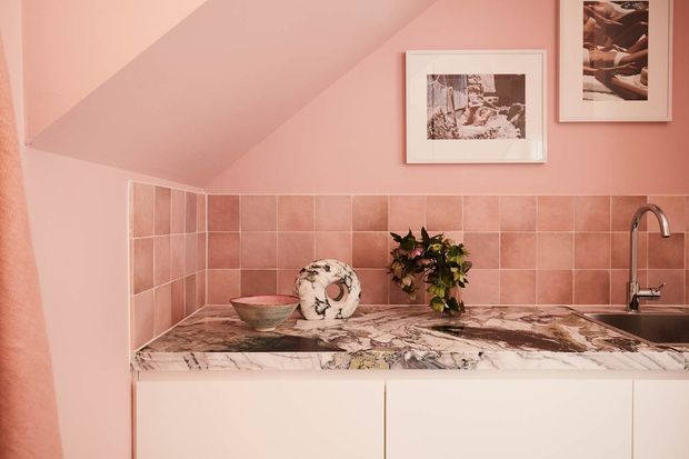 Compact pink-toned kitchen or kitchenette beneath a sloped ceiling, featuring dusty-rose square wall tiles, a dramatic veined marble benchtop, pale flat-front cabinetry, and a brushed metal tap. Framed artwork and decorative objects add a curated, warm character.