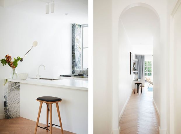 Bright, refined residential interior showing a white kitchen with a slim island or peninsula, marble feature panel, undermount sink and chrome tap, alongside a long white hallway with a tall arched opening and herringbone timber flooring. The hallway leads toward a small furnished room and glazed doors at the rear.