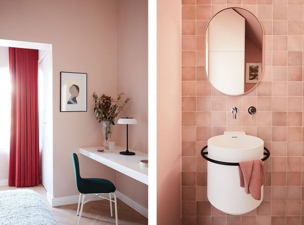A split-view interior image showing a blush-pink bedroom workspace on the left and a compact pink-tiled bathroom vanity on the right. The workspace includes a white built-in desk, dark teal chair, black desk lamp, framed artwork, and red curtains. The bathroom features a wall-mounted white basin with black circular supports, brushed metal fittings, a rounded capsule-shaped mirror, and dusty pink towels.
