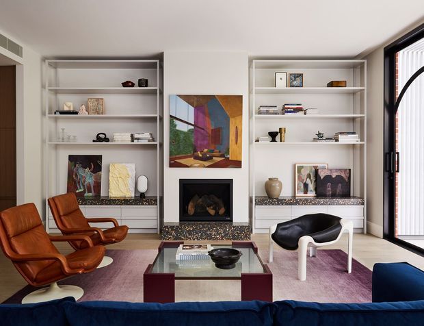 A stylish living room with built-in white shelving flanking a central fireplace, displaying books, ceramics, and artwork. The room includes mid-century leather lounge chairs, a sculptural black-and-white armchair, a glass coffee table, a large blue sofa, colorful artwork, and a dark-framed glazed door or window with vertical blinds. The palette is warm neutral with rich brown, blue, burgundy, and multicoloured accents.