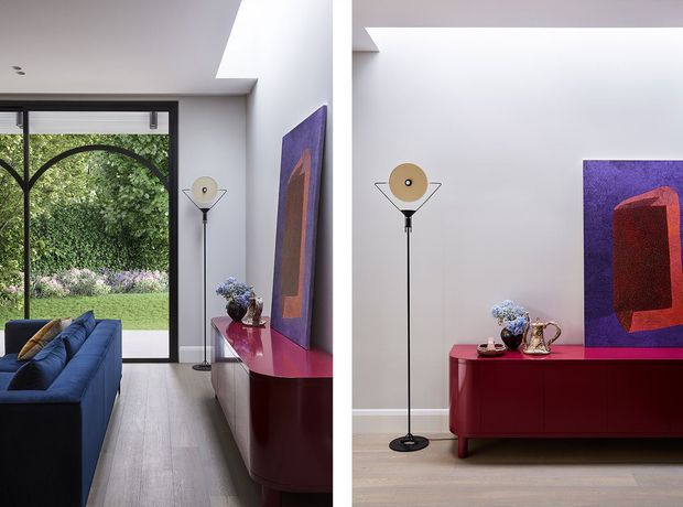 Contemporary living room shown in a split view, featuring a deep blue sofa, pale timber flooring, white walls, a slim black floor lamp, and a glossy burgundy-red sideboard topped with decorative objects. A large purple and red abstract artwork provides a strong colour accent. Full-height glazing opens the room visually to a landscaped garden, and a linear ceiling skylight adds natural light.