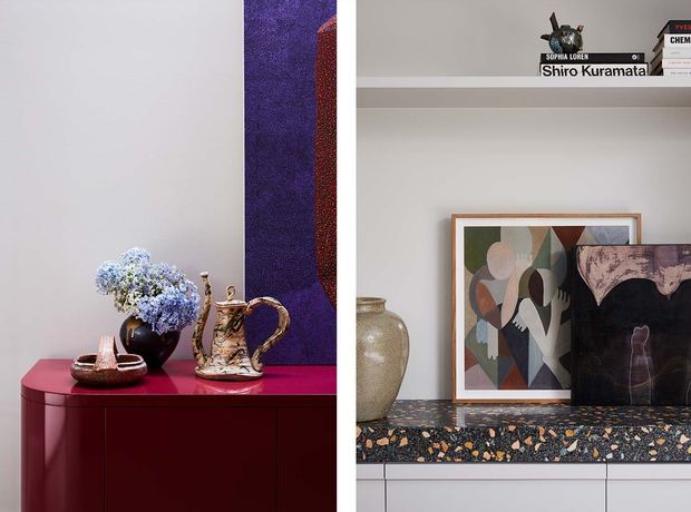 A split interior vignette featuring richly styled display surfaces. On the left, a deep burgundy-red curved-front cabinet supports a dark vase of pale blue flowers, a decorative teapot, and ceramics, with a tall purple artwork behind. On the right, a pale shelving and cabinetry composition displays books, ceramics, framed abstract artwork, and a dark sculptural object above a striking dark terrazzo benchtop with warm stone chips. The overall look is refined, eclectic, and colour-forward.