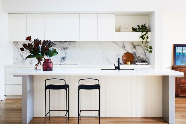 Bright contemporary kitchen with a long white island, vertical-panel cabinetry, two black bar stools, and a dramatic white marble-look splashback. Full-height white overhead cabinetry, open shelving with plants and decor, integrated appliances, timber flooring, and a warm wood-toned adjoining space are visible.