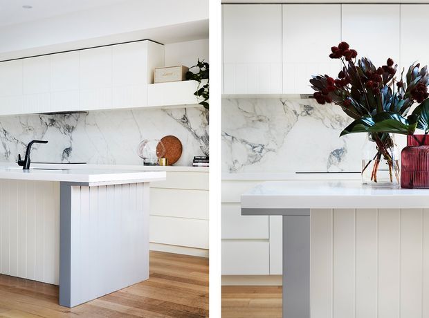 Bright contemporary white kitchen featuring a marble-look splashback, white flat-panel cabinetry, a white island with vertical panel detailing, dark tapware, and warm timber flooring. Decorative flowers and accessories add a small burgundy and green accent.
