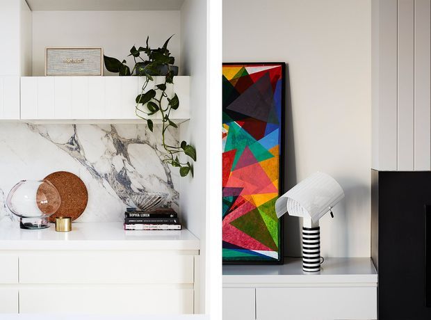 A close-up view of built-in white cabinetry and open display shelving. The left niche features a dramatic white-and-grey marble-look wall finish, books, decorative objects and a trailing plant, while the right niche displays a colourful geometric artwork and a sculptural black-and-white table lamp. The overall composition is clean, contemporary and highly curated.