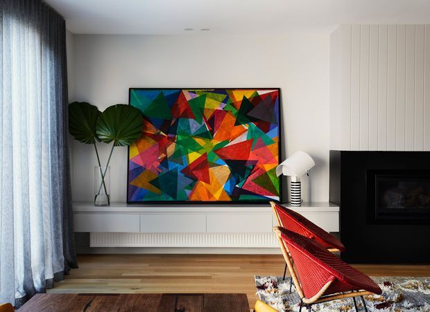 A contemporary living room with a colourful geometric artwork mounted above a long white built-in media cabinet. The room features pale walls, vertical white wall panelling around a black fireplace, full-height sheer and dark curtains, timber flooring, a sculptural floor lamp, and a red woven lounge chair. A patterned rug and timber coffee table add warmth and texture.