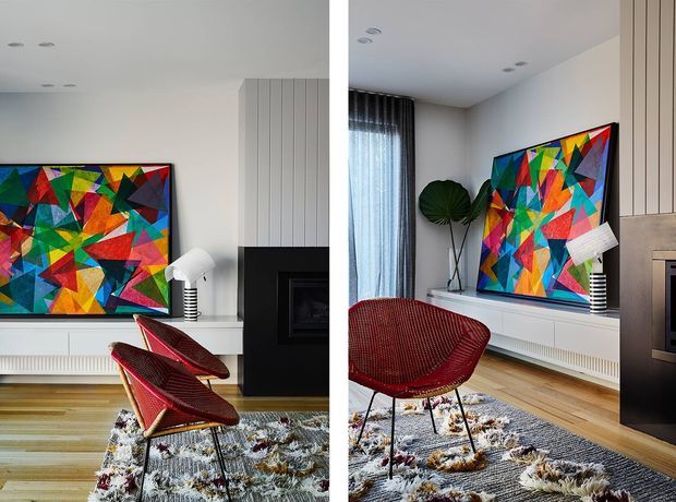 A bright, contemporary living room shown from two similar angles, featuring a large colourful geometric artwork above a low white media console, a black fireplace surround, vertical wall panelling, timber flooring, a textured area rug, and red woven lounge chairs. Floor-to-ceiling curtains, a sculptural floor plant, and recessed ceiling lights complete the room.