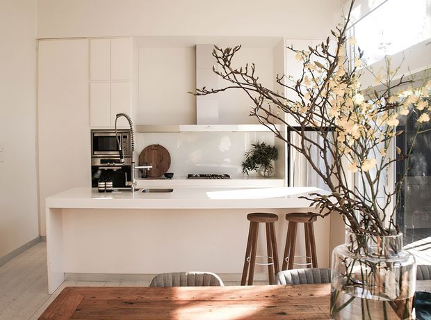 Bright contemporary open-plan kitchen and dining area with a large white island, integrated cabinetry, stainless-steel appliances, pale splashback, timber bar stools and a timber dining table. Tall glazing brings in abundant natural light, while a large branch arrangement forms a prominent decorative feature in the foreground.