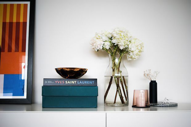 A styled interior console vignette against a white wall, featuring a framed abstract artwork, teal storage boxes, a sculptural bowl, a tall glass vase of white hydrangeas, a black vase with delicate branches, a candle holder, and small decorative objects on a minimalist white cabinet.
