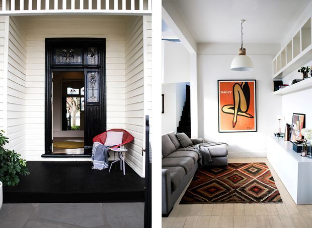 Split-view image showing a traditional home entry facade on the left and a bright contemporary living room on the right. The exterior features white horizontal weatherboard cladding, a black-painted raised entry platform, and an ornate black glazed front door beneath a white balustrade. The interior has white walls, pale timber flooring, a grey sectional sofa, a geometric patterned rug, white built-in shelving and cabinetry, a large framed artwork, and a pendant light.