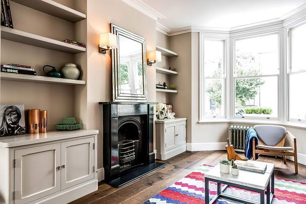 A refined traditional living room with a black marble fireplace and mantel as the focal point, built-in open shelving and painted cabinetry, a large bay window, and a mid-century-style lounge chair. The room combines warm taupe walls, white architectural trim, dark timber flooring, and a colourful patterned rug.