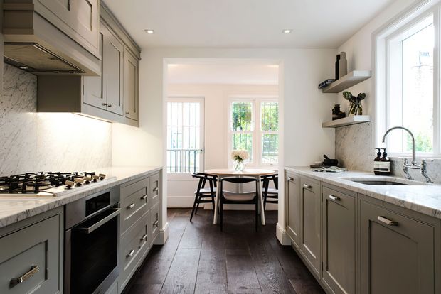 A bright, refined galley kitchen with grey-green shaker cabinetry, white marble-look countertops and splashbacks, integrated appliances, and a dark timber floor. A dining area with a round table and chairs sits beyond the kitchen, with abundant natural light from tall windows and glazed doors.