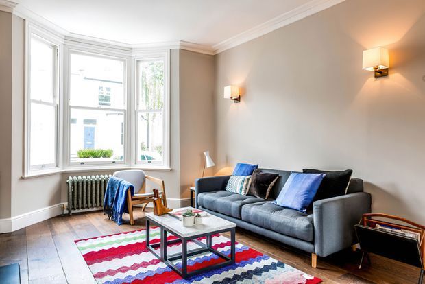 A bright, traditionally detailed living room with a grey sectional sofa, colourful patterned rug, timber coffee table, lounge chair, and a vintage-style side table. The room features large bay windows, white-painted trim and cornicing, warm taupe walls, timber flooring, wall sconces, and a visible cast-iron radiator.
