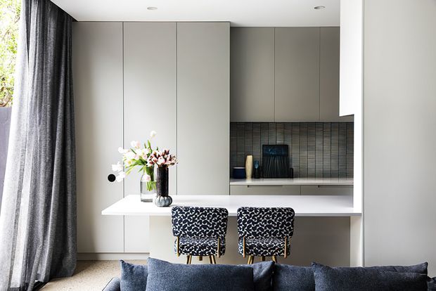 A refined open-plan living area with a compact contemporary kitchen behind a white peninsula island. Full-height pale cabinetry, a dark vertically textured tiled splashback, integrated appliances, sculptural pendant-free recessed lighting, and patterned bar stools define the kitchen. A dark sofa, full-height charcoal curtains, and floral arrangement occupy the foreground, with abundant daylight entering from the left.